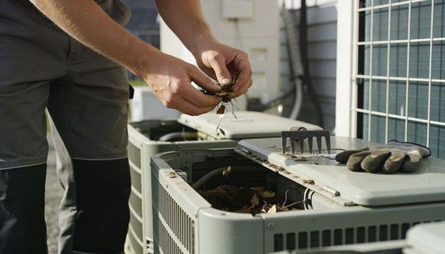 Hvac technician clearing leaves from outdoor air conditioner condenser during maintenance