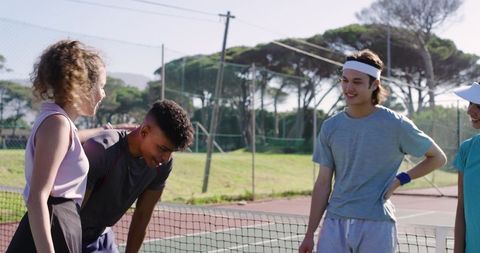Friends laughing and catching breath at net after match on sunny outdoor tennis court