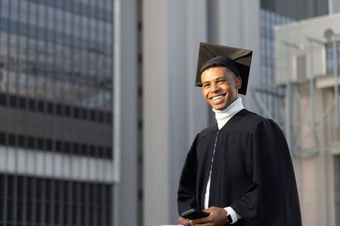 African american graduate smiling in cap and gown with smartphone by urban glass facades