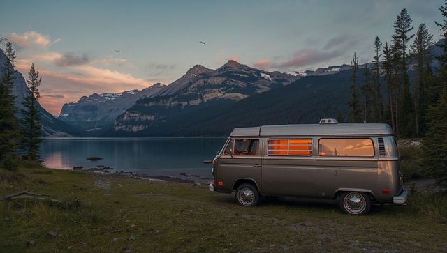 Vintage camper van at mountain lake sunset with warm interior glow and alpine reflection