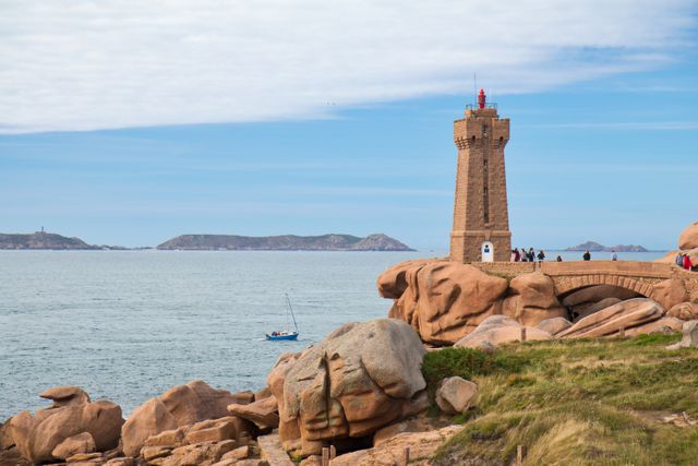 This image shows the scenic beauty of a lighthouse standing majestically on rocky shoreline of Ploumanac'h, France. Background includes calm ocean with sailboat. Ideal for travel websites, nature blogs, tourism promotions, and inspirational landscape prints.