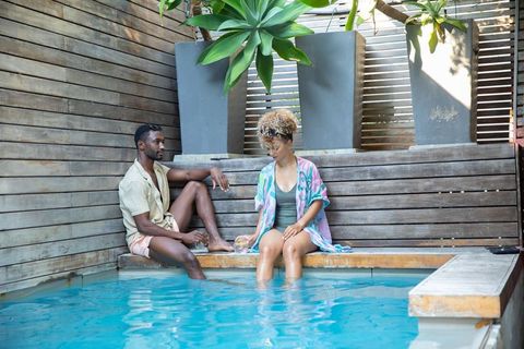 Diverse couple relaxing poolside on wooden bench dipping feet in turquoise pool