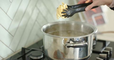 Woman lowering ribbon pasta into boiling stainless steel pot with tongs on gas stove