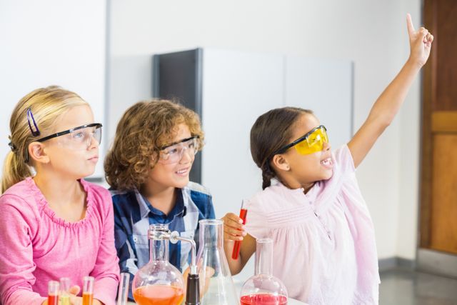 Children are conducting a science experiment in a school laboratory, wearing safety goggles and working with beakers and chemical solutions. One child is raising their hand enthusiastically. This image can be used for educational materials, school promotions, science program advertisements, and STEM-related content.