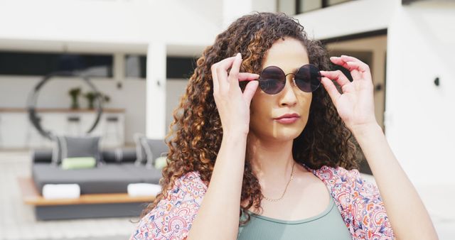 Fashion-forward woman with curly hair adjusting sunglasses, outdoor luxurious setting. Ideal for advertising summer fashion, eyewear brands, or articles on style and confident living.