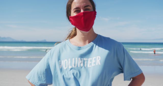 Person stands on a beach wearing a blue volunteer shirt and red face mask. The clear blue ocean and a bright sky are in the background. This image is ideal for illustrating community service, volunteering efforts, outdoor activities, beach clean-ups, and social conscious movements.