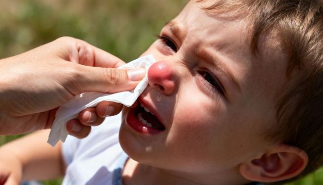 Crying toddler having nose gently wiped by caregiver outdoors in sunlight, parental care