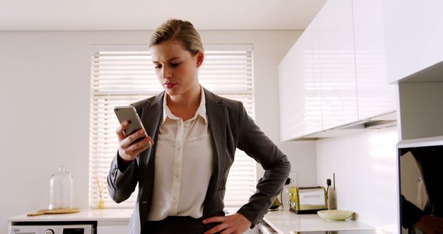 Businesswoman focusing on smartphone, reading a text message during her morning in a modern kitchen. This could be used for concepts like work-life balance, importance of morning routines, professional communication, and modern lifestyles.