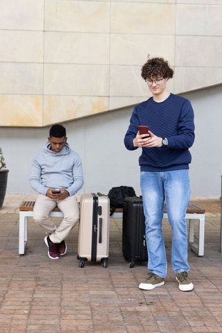 Multiracial young men checking phones while waiting with suitcases at urban plaza