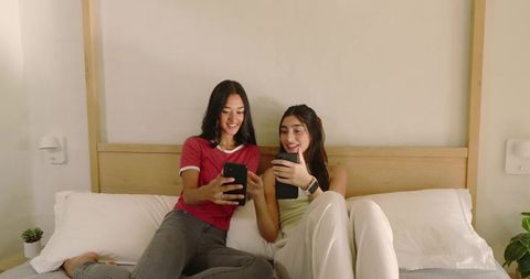 Diverse Female Friends Sharing Screens Relaxing on Bed, Genuine Smiles and Connection