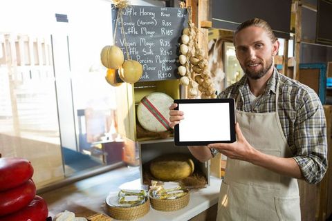 Male Gourmet Shopkeeper Presenting Tablet in Rustic Cheese Shop