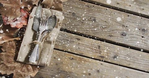 Rustic autumn table scene with cutlery on weathered wood, napkin tied with twine, falling snow