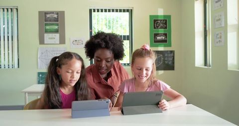 African American Teacher Guiding Two Elementary Girls Using Tablets in Bright Classroom