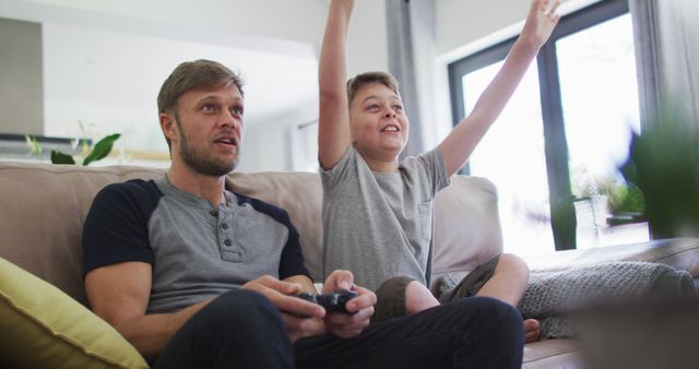 Father and son enjoying a video game together while sitting on a couch in living room. The son is cheerfully raising his hands in excitement while father is focused on the game. Perfect for depicting quality family time, gaming experiences, and parent-child bonding moments. Suitable for advertisements, lifestyle blogs, and publications about family activities and relationships.