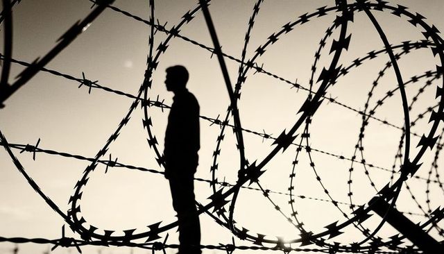 Standing silhouette of man framed by coils of razor wire on perimeter fence at sunset