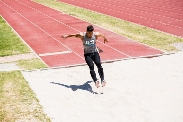Athlete in mid-air performing a long jump during a track and field competition. The athlete is wearing a number bib and landing in the sand pit. Ideal for use in sports articles, fitness blogs, athletic training materials, and promotional content for sporting events.