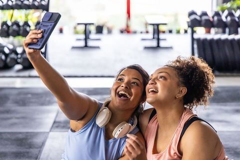 Two diverse women laughing while taking selfie after workout in sunlit gym