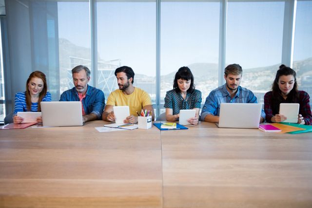 Creative team of professionals sitting in a row at a large wooden table, actively working on laptops and tablets in a modern office. Ideal for illustrating teamwork, collaborative work environments, modern technology usage, and professional settings. Useful for business-related content, office productivity themes, and digital collaboration visuals.