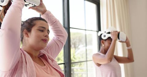 African american friends trying virtual reality headsets at home wearing pink casual tops