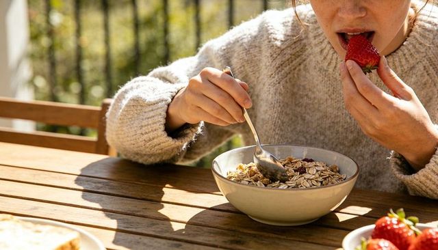 Woman eating strawberry with granola bowl outdoors on sunlit wooden table breakfast