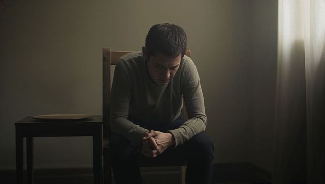 Man leaning forward contemplating on chair by window in dim minimalist interior