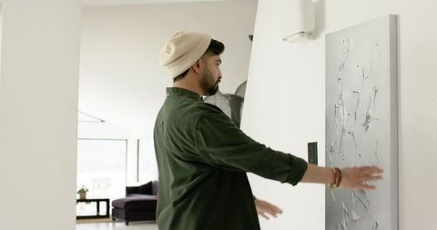 Indian man hanging minimalist textured grey canvas in modern bright hallway wearing beanie