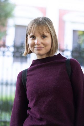 Smiling young woman wearing maroon sweater and backpack posing on urban street with bokeh