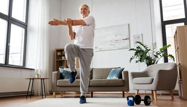 Senior man doing balance stretch and home workout with kettlebell in sunlit living room