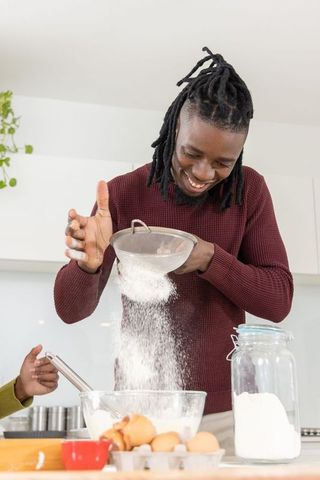 African american man smiling while sifting flour for baking with helper in modern kitchen