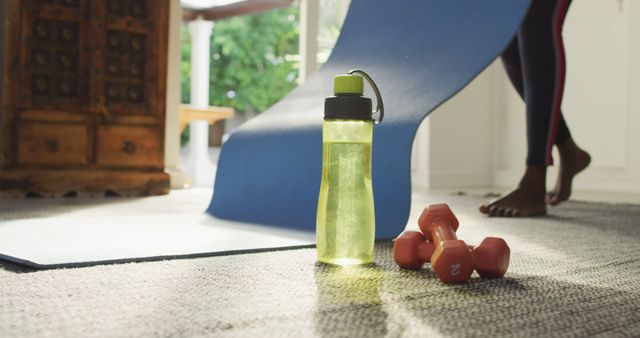 Water bottle, dumbbells, and yoga mat with woman preparing for workout. Ideal for promoting home fitness routines, healthy lifestyles, and indoor exercises. Can be used in articles or blogs about staying fit at home.