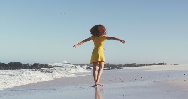 Woman walking barefoot on sandy beach with arms open, wearing yellow dress and enjoying the sea breeze. Perfect for promoting relaxation, summer vacations, freedom, nature retreats, and coastal lifestyle advertisements.
