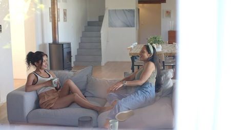 Two diverse women laughing and chatting, sipping mugs on gray sectional sofa in sunlit living room