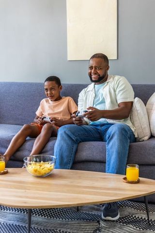 African american father and son playing video games on gray sofa with snacks and juice