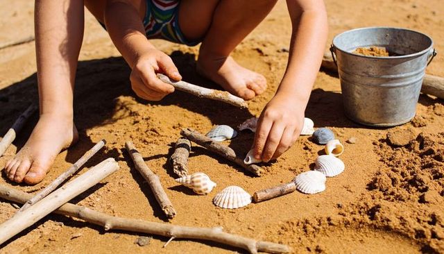 Child arranging seashell and driftwood circle on sunny beach sand with metal bucket