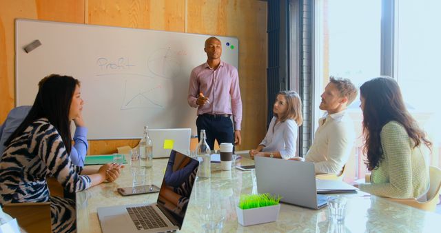 Business professionals are gathered in office for important presentation led by male colleague at whiteboard discussing profit. Laptops and documents are on table, emphasizing strategic planning and teamwork. Useful for illustrating corporate training, business strategy discussions, or teamwork in office settings.