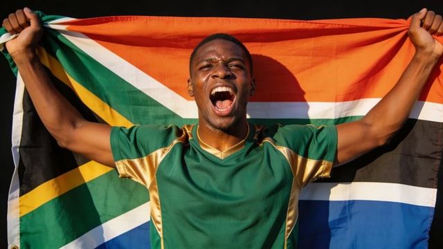 Triumphant athlete shouting while holding south african flag wearing green and gold jersey