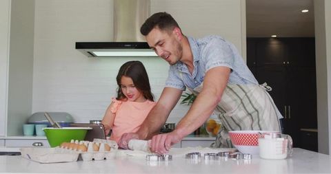 Smiling father and daughter baking and rolling dough using tablet recipe on kitchen island