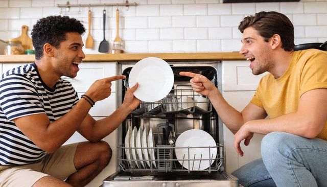 Smiling friends loading dishwasher together in bright modern kitchen, casual lifestyle