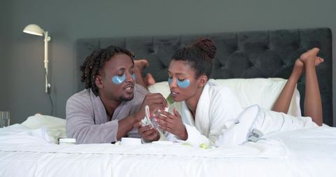Couple relaxing with under-eye gel patches on bed in bathrobes enjoying skincare ritual