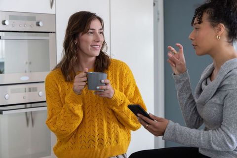 Two women chatting in modern kitchen, mustard sweater holding mug, casual conversation