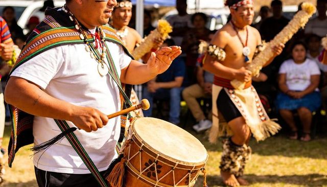 Indigenous drummer leading traditional celebration, wearing woven shawl and playing drum
