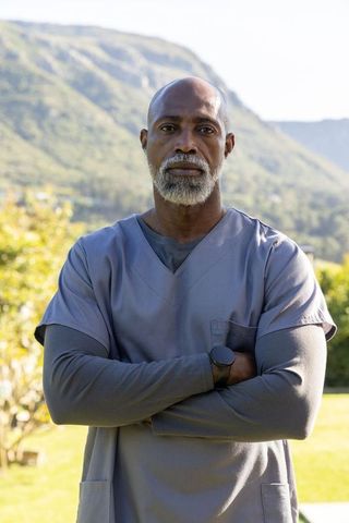 Senior african american nurse standing arms crossed wearing gray scrubs in sunlit yard