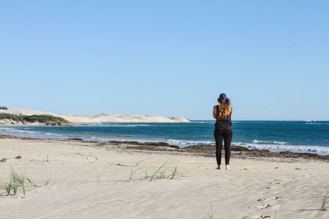 Woman is standing on a sandy beach facing the sea, wearing black clothing and a blue hat. The ocean has gentle waves, and the sky is clear. The setting includes sand dunes in the background. Useful for themes of solitude, relaxation, travel, nature, and outdoor activities.