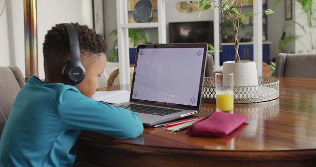 Young boy wearing headphones sitting at a wooden table, using a laptop for online education. Digital education materials, a glass of juice, and stationary items are beside him. The image is ideal for illustrating themes related to remote learning, child education, home schooling, and the use of technology in education.