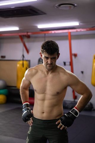 Muscular shirtless fighter standing with hands on hips in gritty combat gym