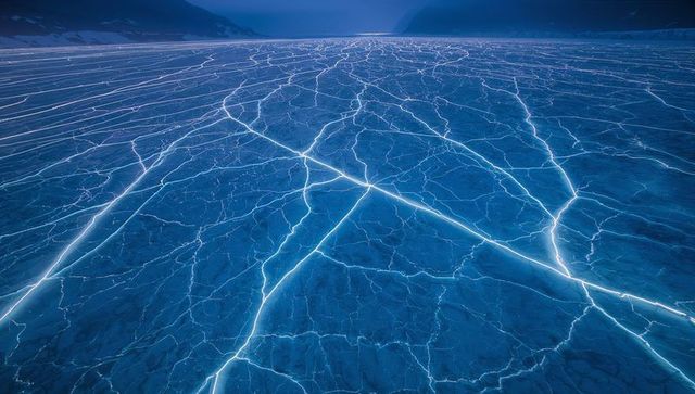 Glowing ice cracks on frozen lake at twilight in remote mountain valley, blue fractal