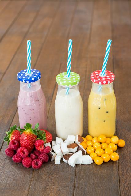 Three bottles filled with colorful smoothies, each topped with a polka dot lid and striped straw, are placed on a wooden table. Fresh strawberries, raspberries, coconut pieces, and yellow berries are arranged in front of the bottles. This vibrant and healthy scene is perfect for promoting nutritious drinks, summer refreshments, or healthy lifestyle content.
