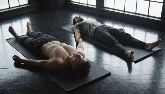 Male partners practicing savasana holding hands in moody backlit yoga studio