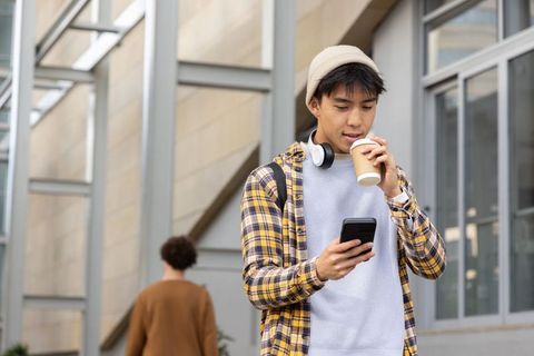 Asian commuter checking smartphone while sipping coffee in modern urban atrium wearing plaid shirt