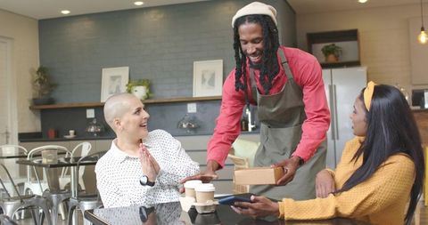 Smiling african american barista serving takeaway box to two women in cozy cafe